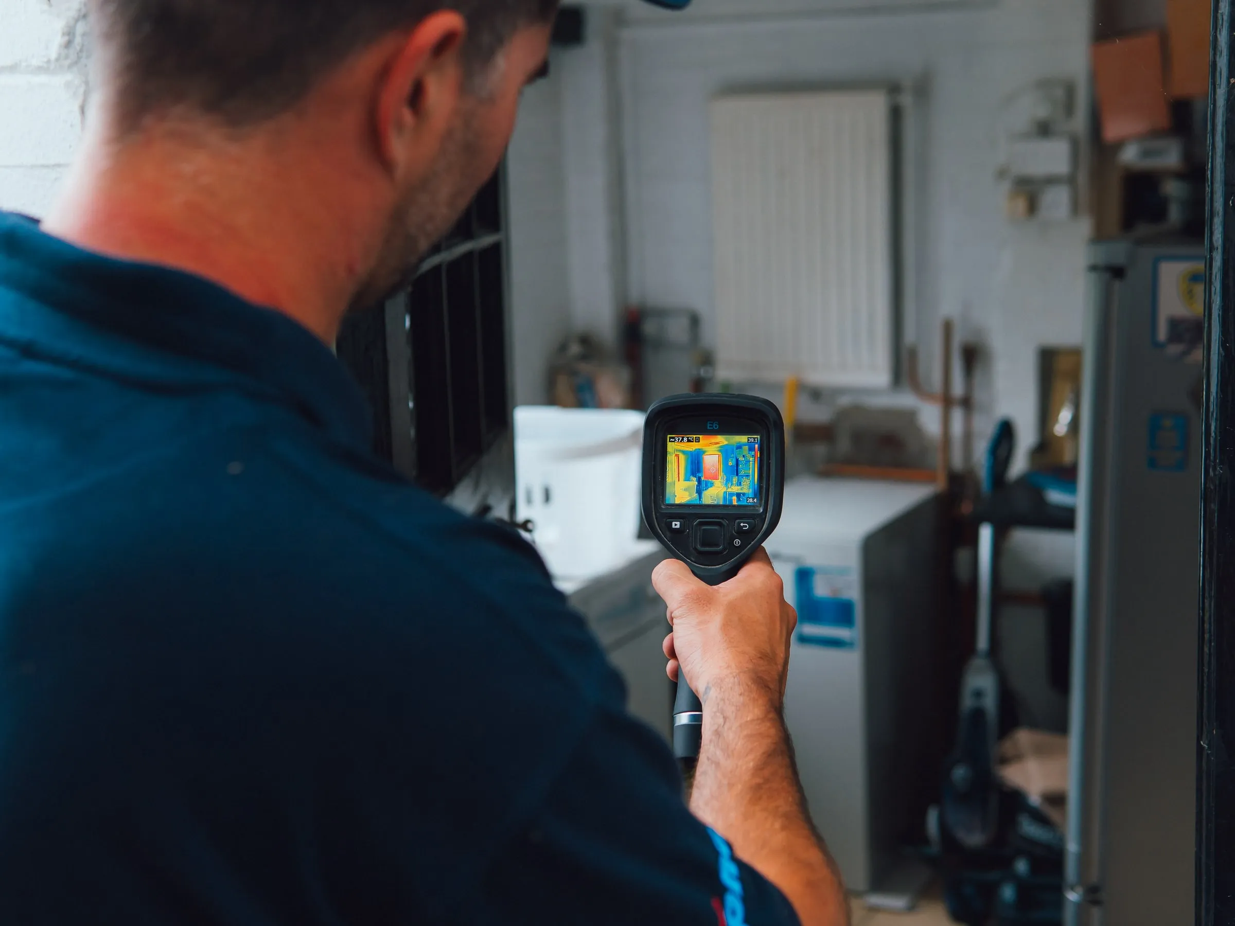 Engineer pointing a thermal camera at a boiler and cylinder in a plant room, the screen showing the heat pattern