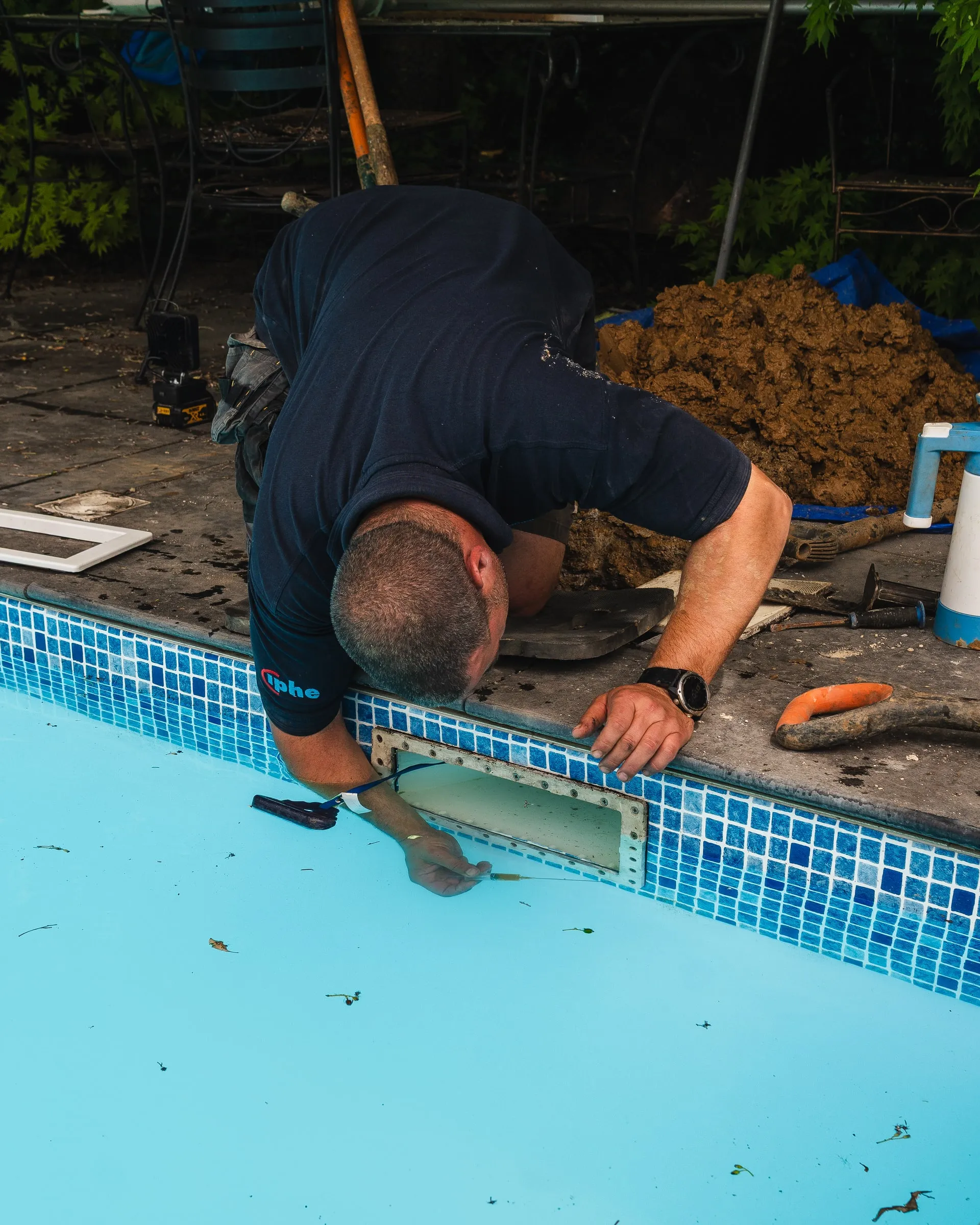 Engineer leaning over the edge of a tiled pool to inspect a fitting in the pool wall